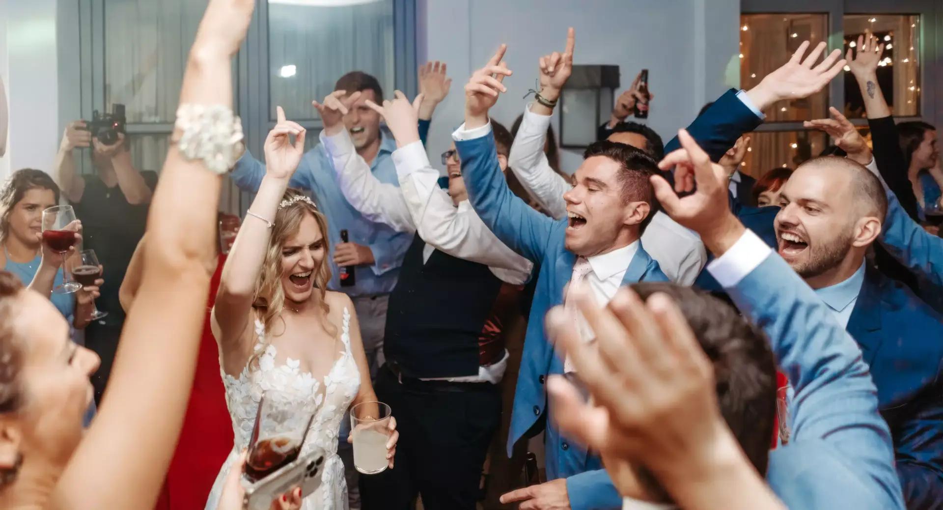 A group of people, including a bride in a white dress, raise their arms and cheer enthusiastically while dancing at a lively indoor celebration.