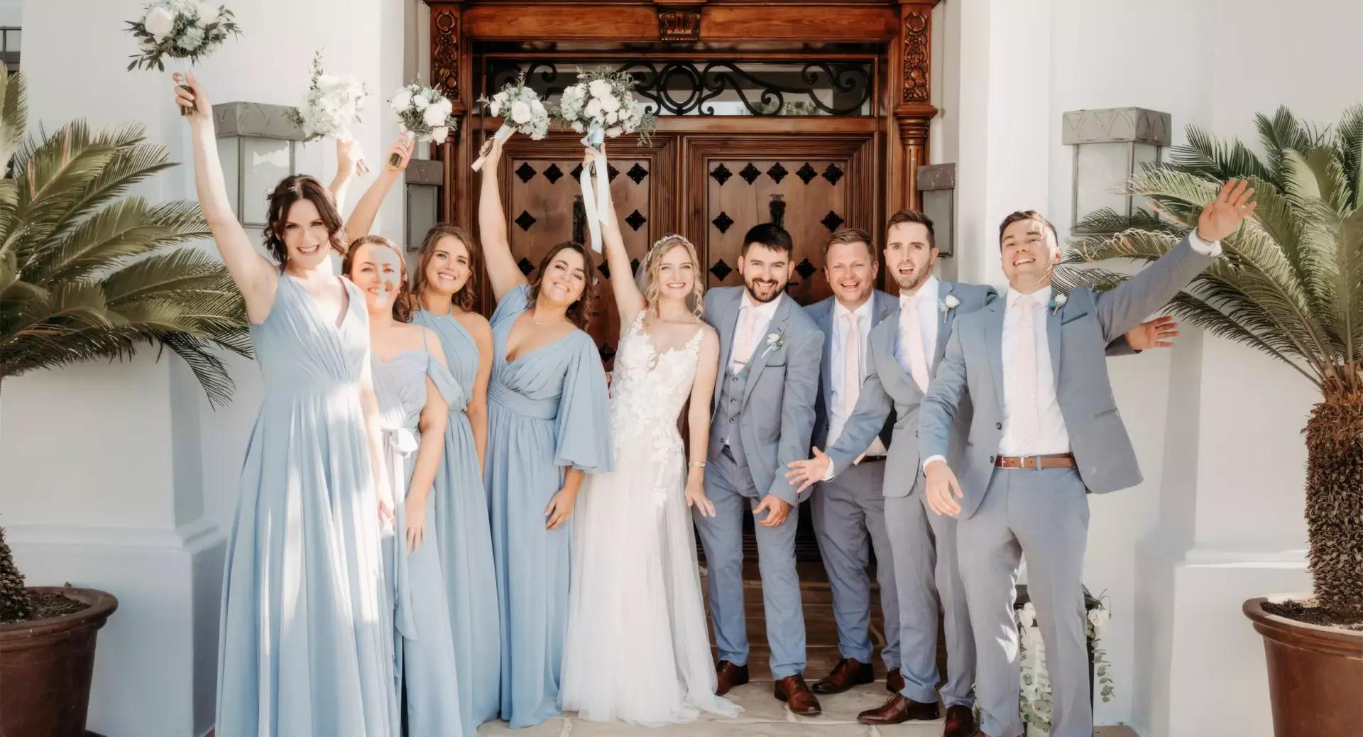 A bride and groom pose with their wedding party, who are dressed in light blue dresses and suits, standing in front of ornate wooden doors and flanked by potted palm plants.