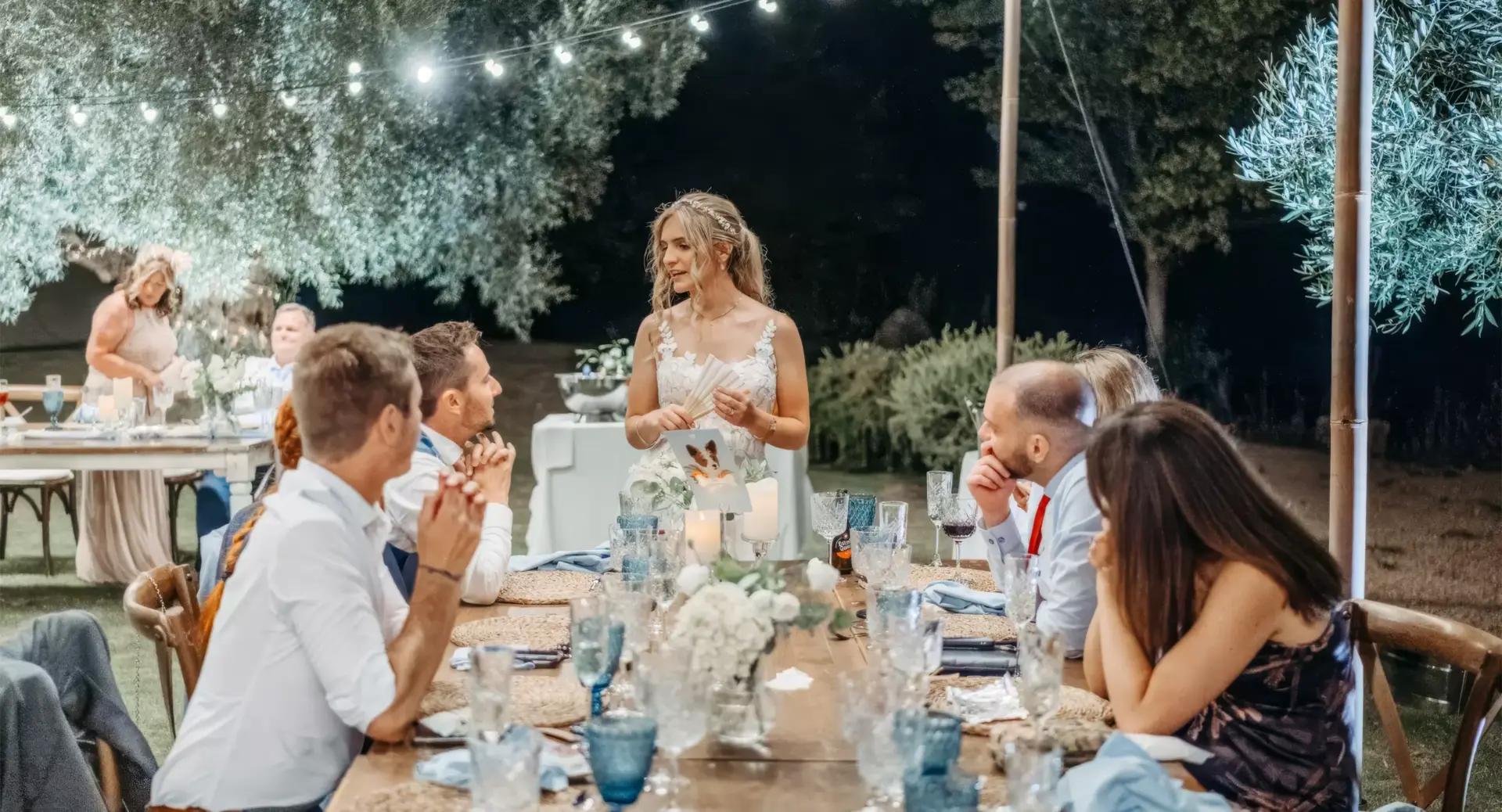 A woman in a white dress stands and speaks at an outdoor dinner table while guests seated around her listen during an evening event.