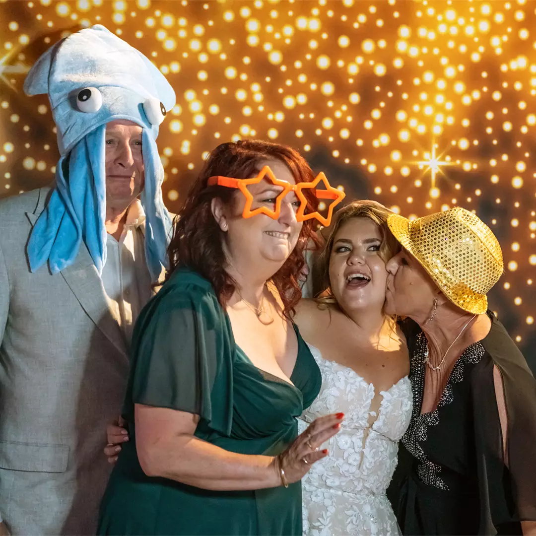 Four adults pose in front of a sparkling backdrop, wearing playful accessories including a squid hat, star-shaped glasses, and a gold hat; one woman whispers to a smiling bride.