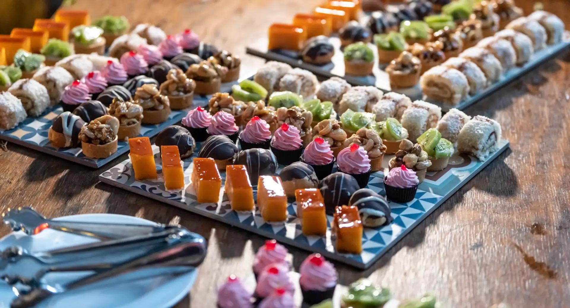 An assortment of bite-sized desserts, including pastries, cakes, and fruit-topped treats, arranged neatly on trays on a wooden table.