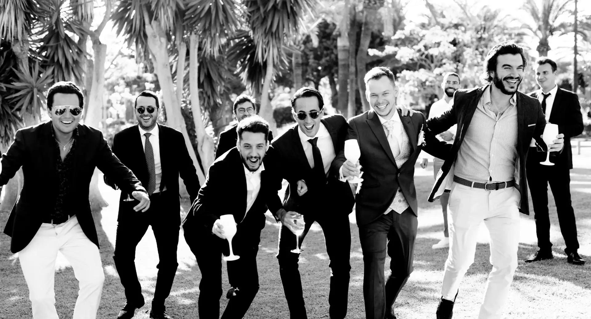 A group of men in formal attire, holding drinks and smiling, walk together outdoors among palm trees in bright sunlight.