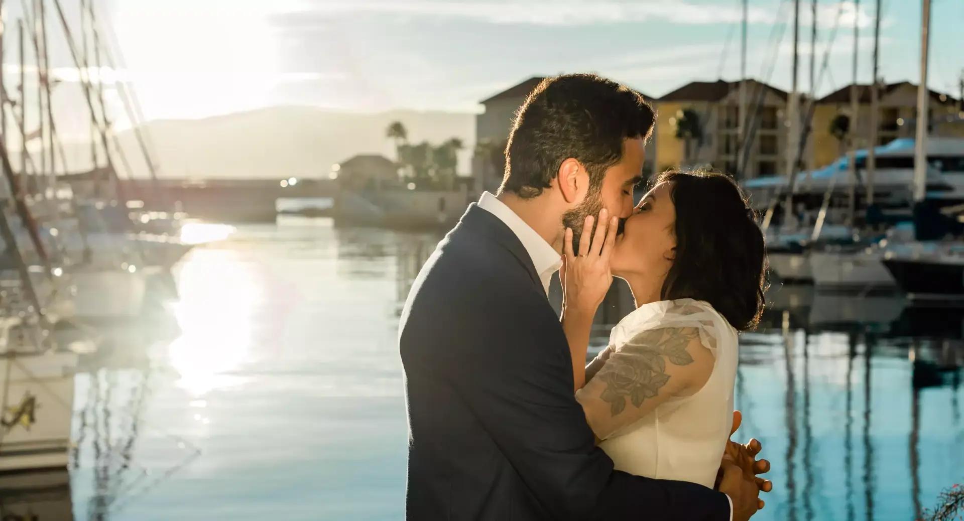 A couple dressed formally shares a kiss by a marina with boats and buildings in the background at sunset.