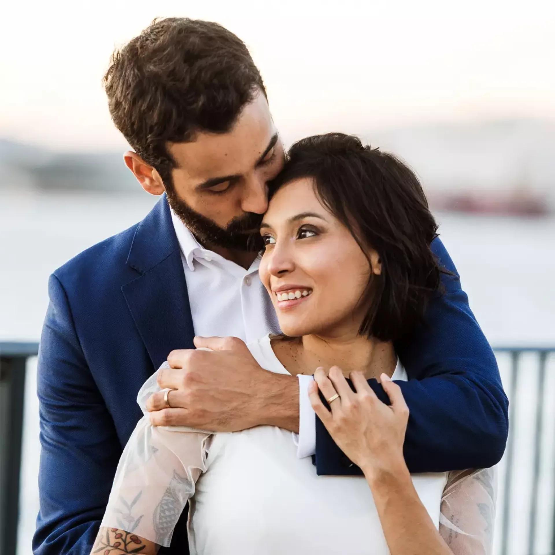 A man in a blue suit hugs a woman in a white dress from behind; he kisses her head while she smiles and looks away.