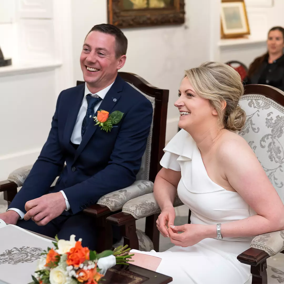 A man in a navy suit and a woman in a white dress sit smiling together at a table with a bouquet of flowers, in a formal indoor setting.