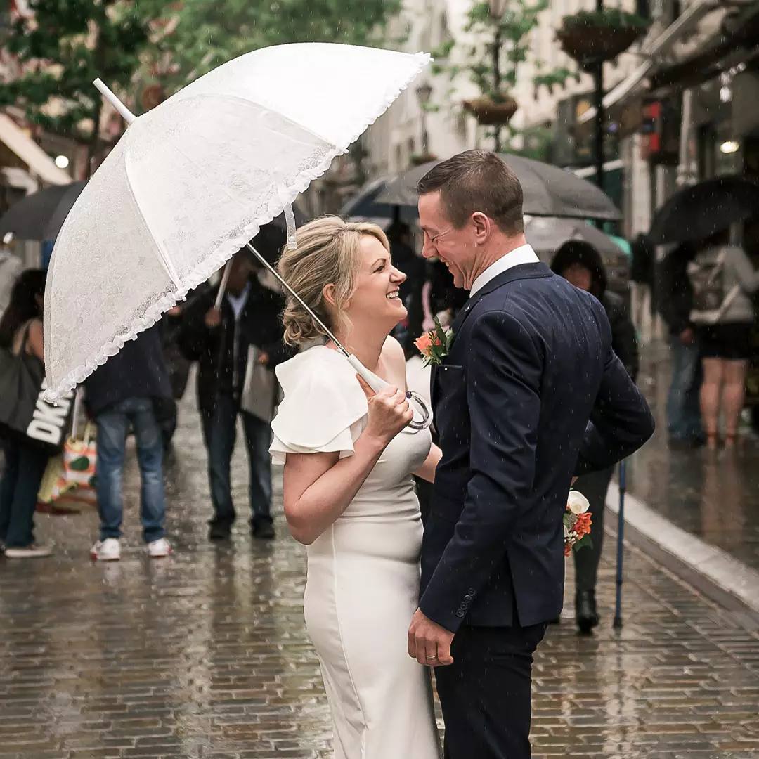 A bride and groom stand close together under a white umbrella on a rainy city street, smiling at each other, with people and umbrellas in the background.