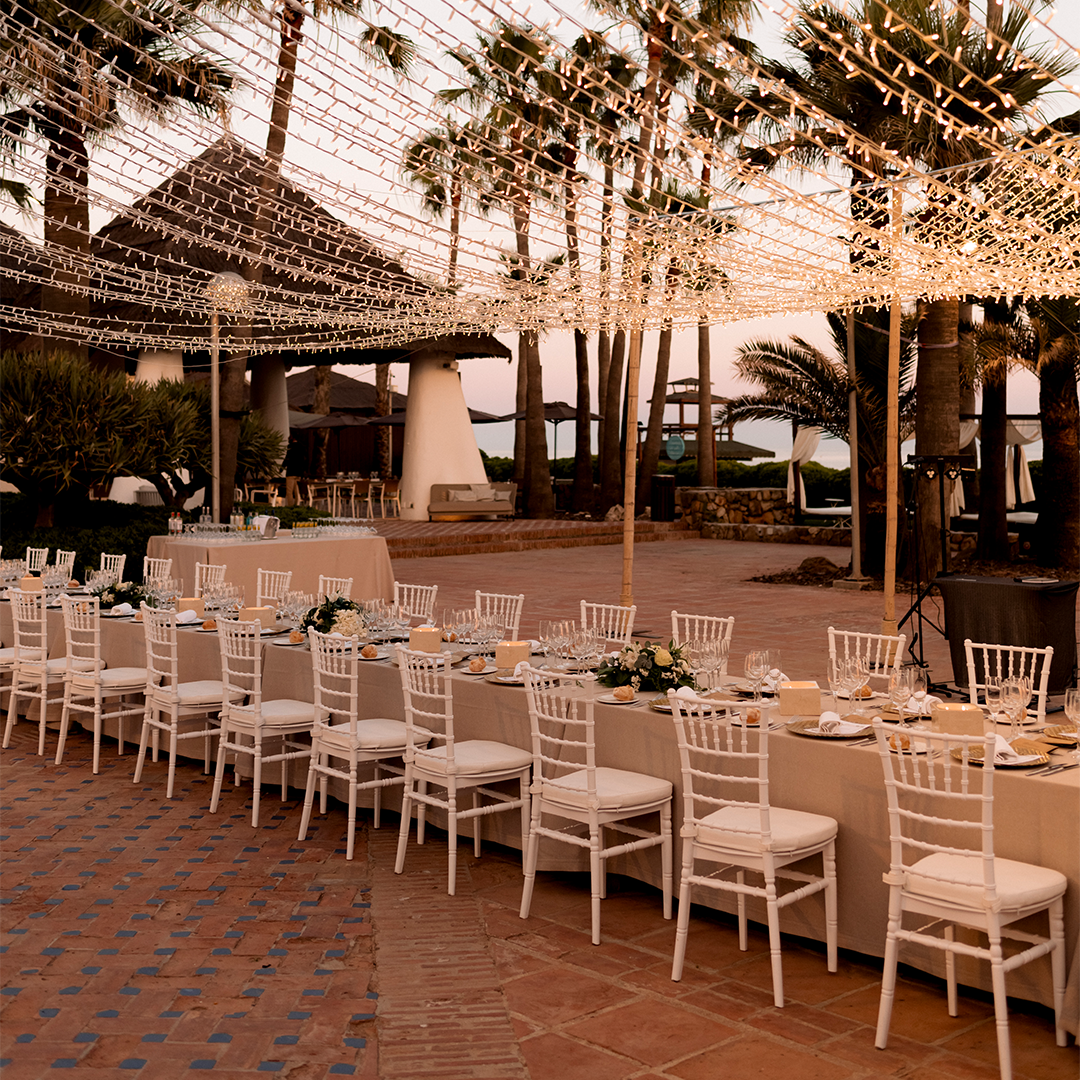 Long banquet tables with white chairs are set for an outdoor event under string lights, with palm trees and a thatched building in the background.