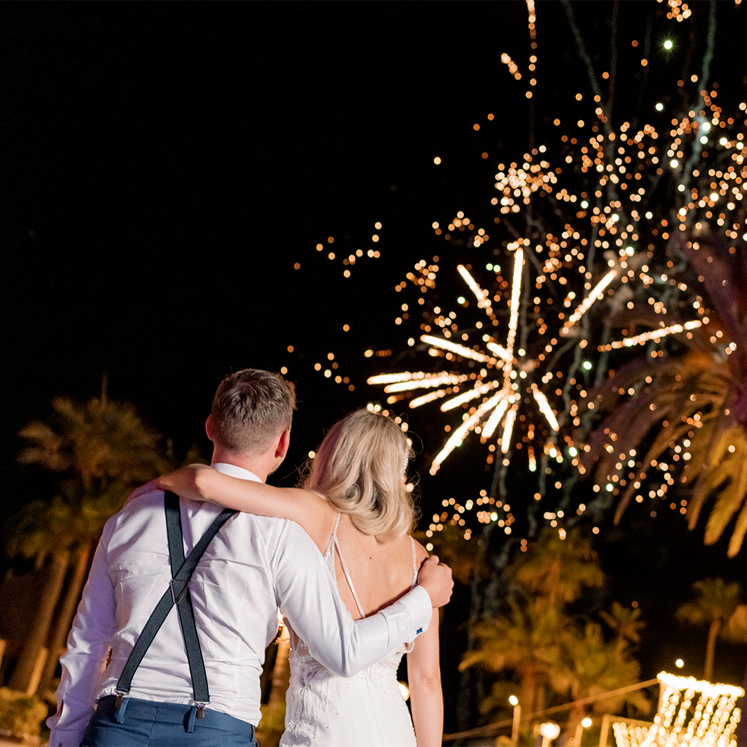 A couple in formal attire stands arm-in-arm, facing away from the camera, watching fireworks light up the night sky.