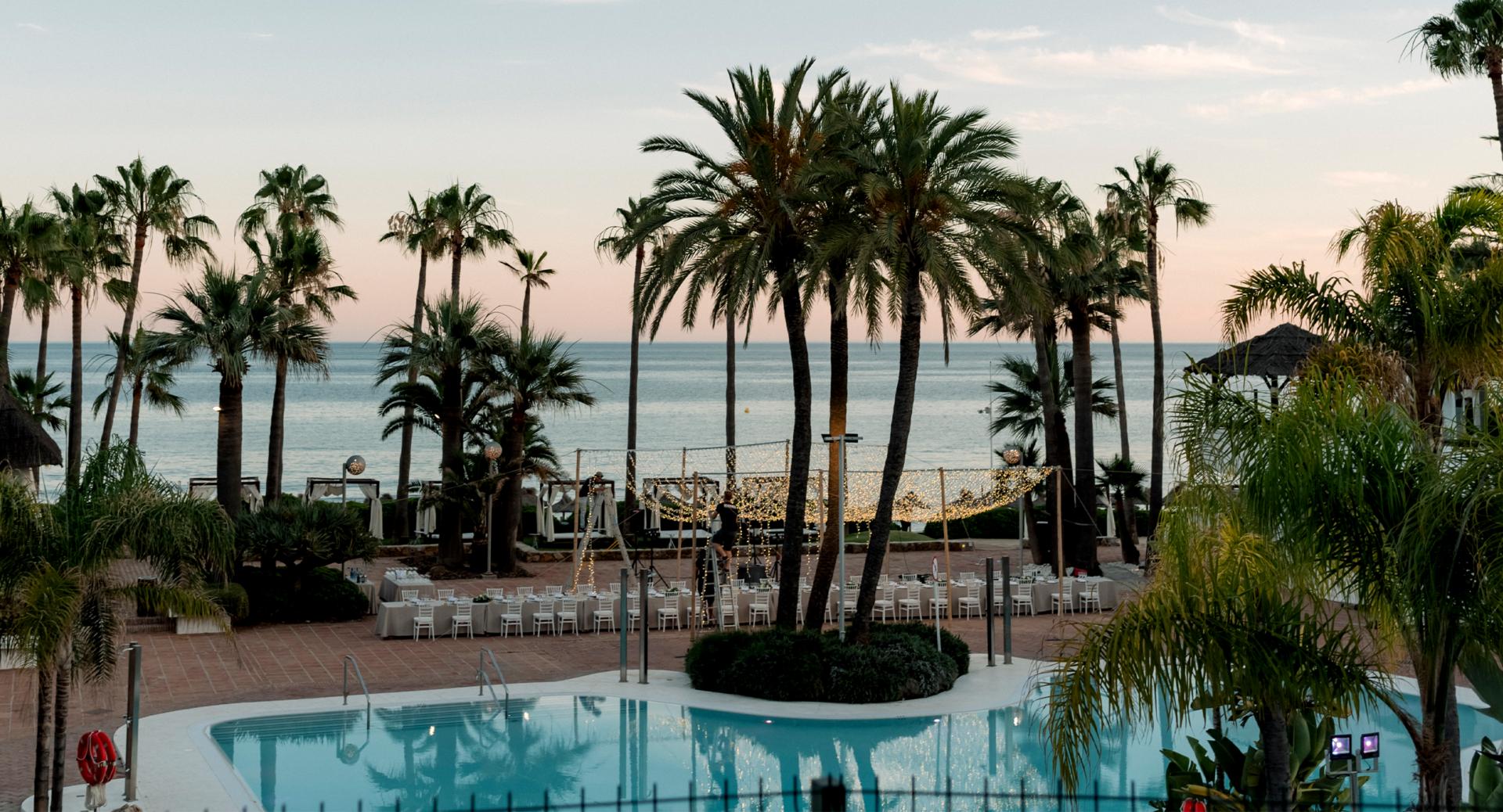 Outdoor pool area with palm trees, rows of white chairs and tables set up, and ocean visible in the background under a cloudy sky at sunset.