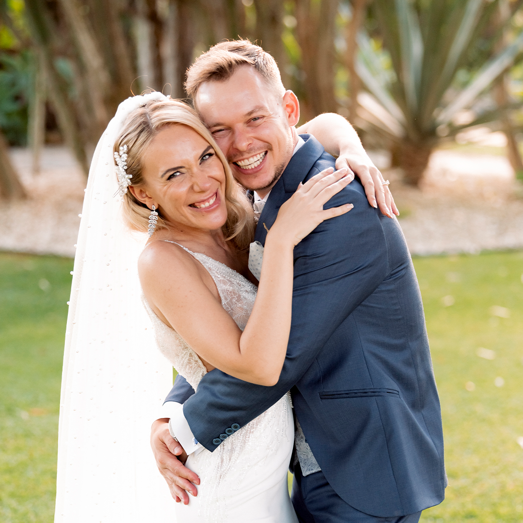 A bride and groom in wedding attire stand outdoors, smiling and hugging closely, with greenery in the background.