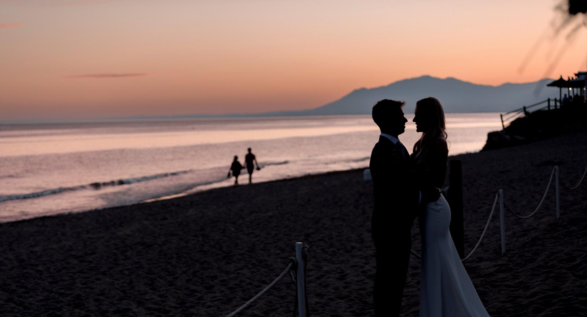 A couple stands closely together on a beach at sunset, with another couple walking by the shoreline in the background.