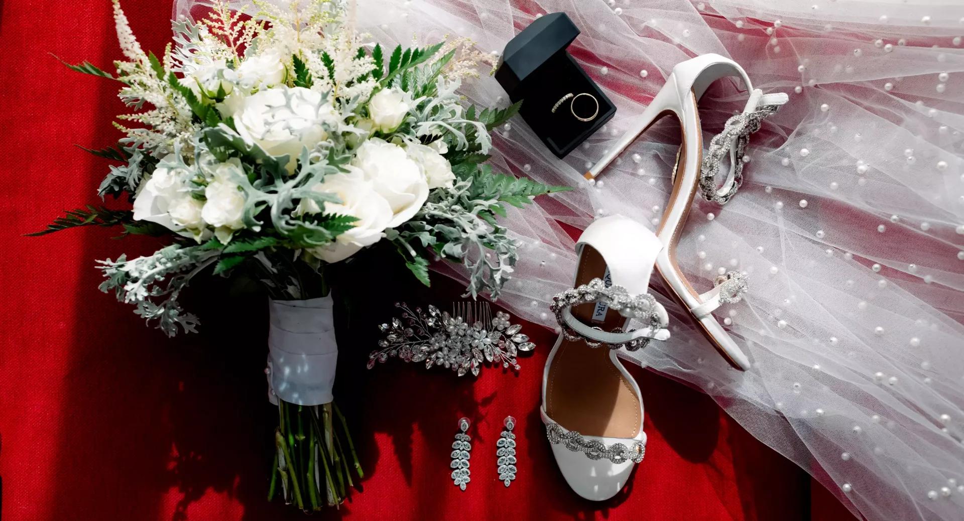 A bridal bouquet, white high-heeled shoes, jewelry, a ring in a box, and a pearl-embellished veil are arranged on a red surface.