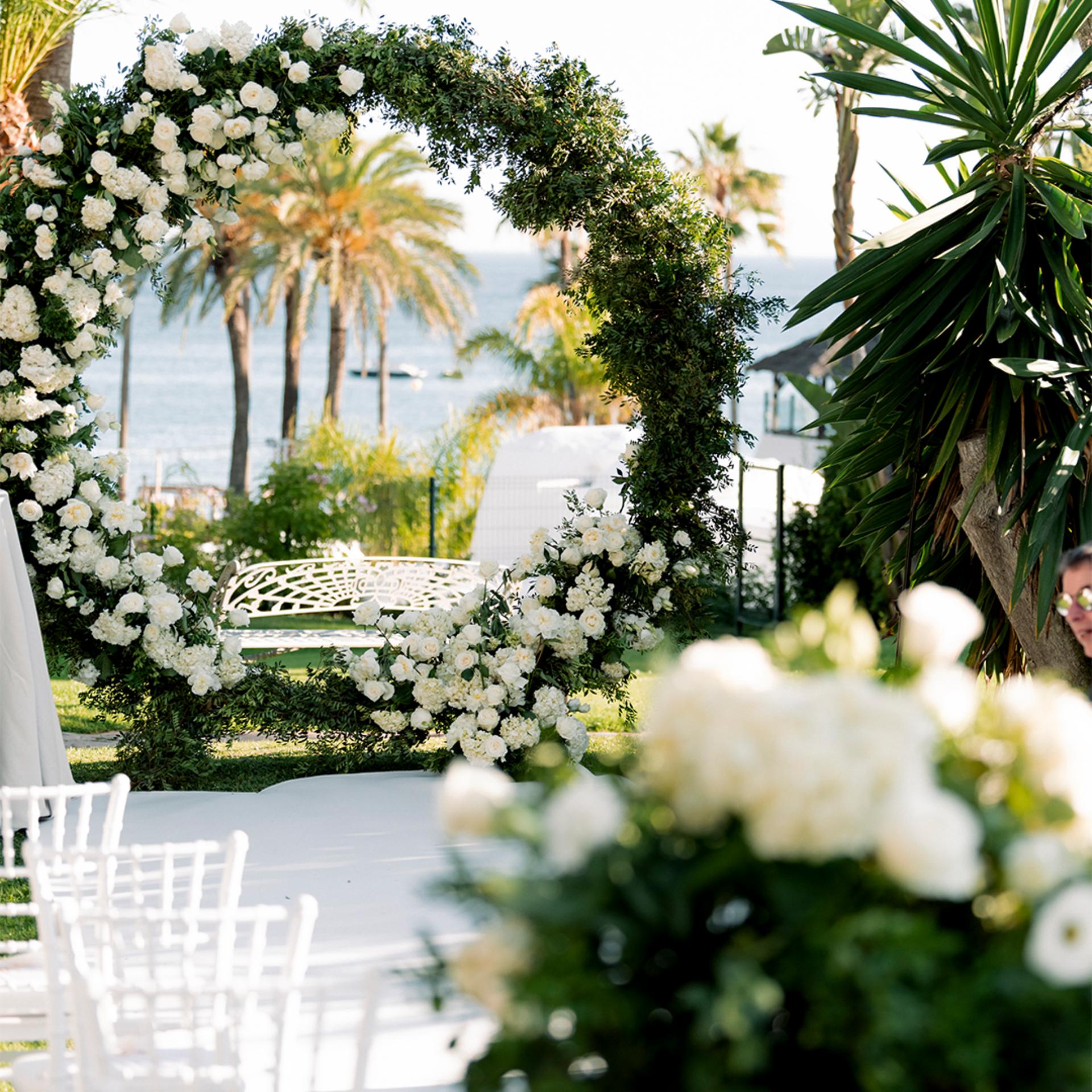 A circular floral arch decorated with white flowers and greenery stands near white chairs at an outdoor wedding venue by the water, with palm trees in the background.