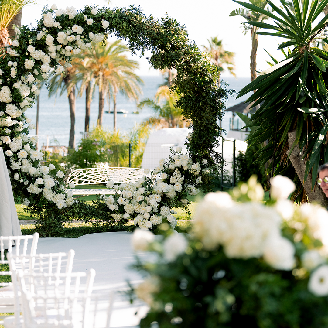 A circular floral arch decorated with white flowers and greenery stands near white chairs at an outdoor wedding venue by the water, with palm trees in the background.