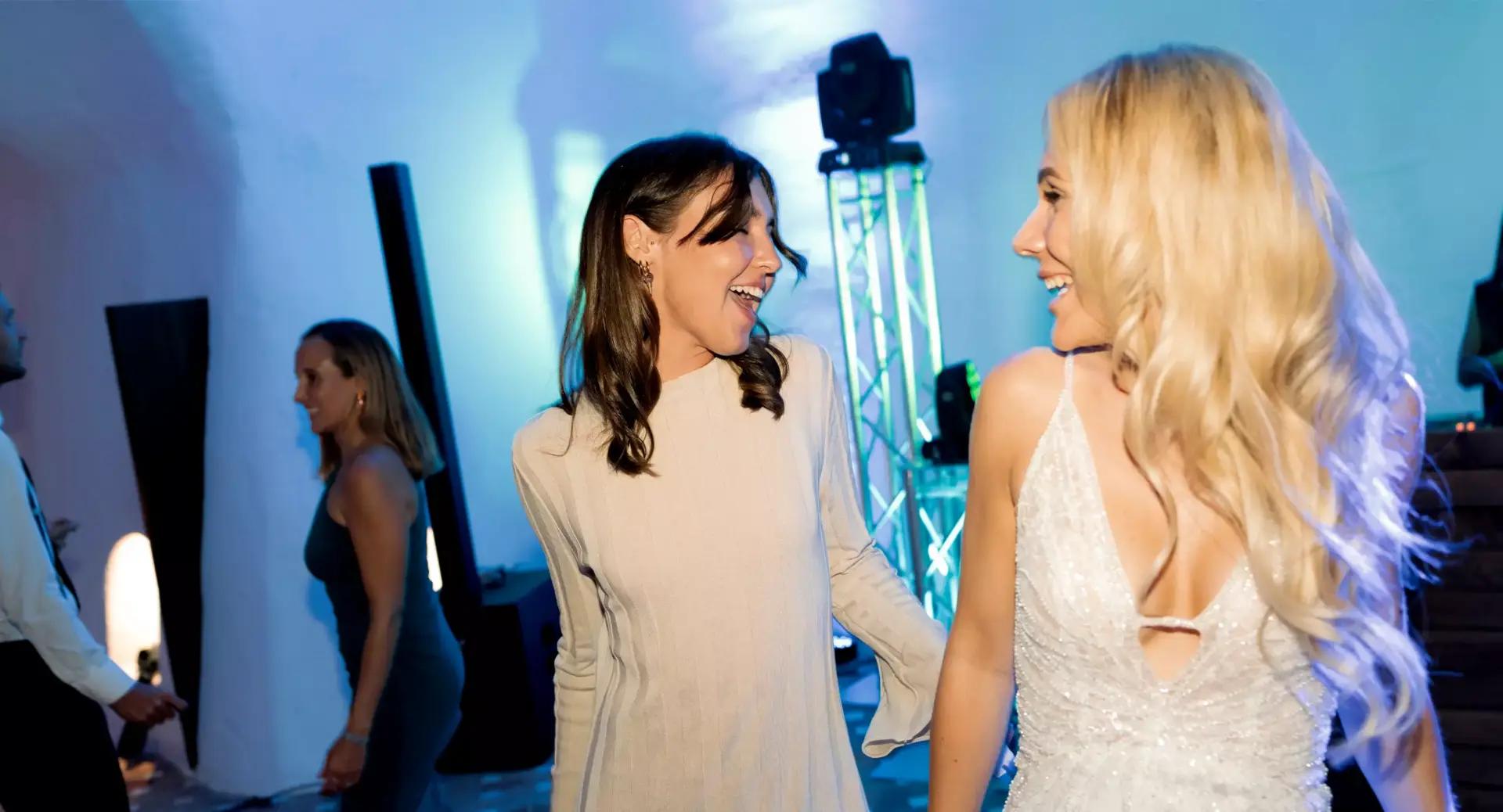 Two women, one in a light dress and one in a white gown, smile and hold hands while dancing at an indoor event with colorful lighting.