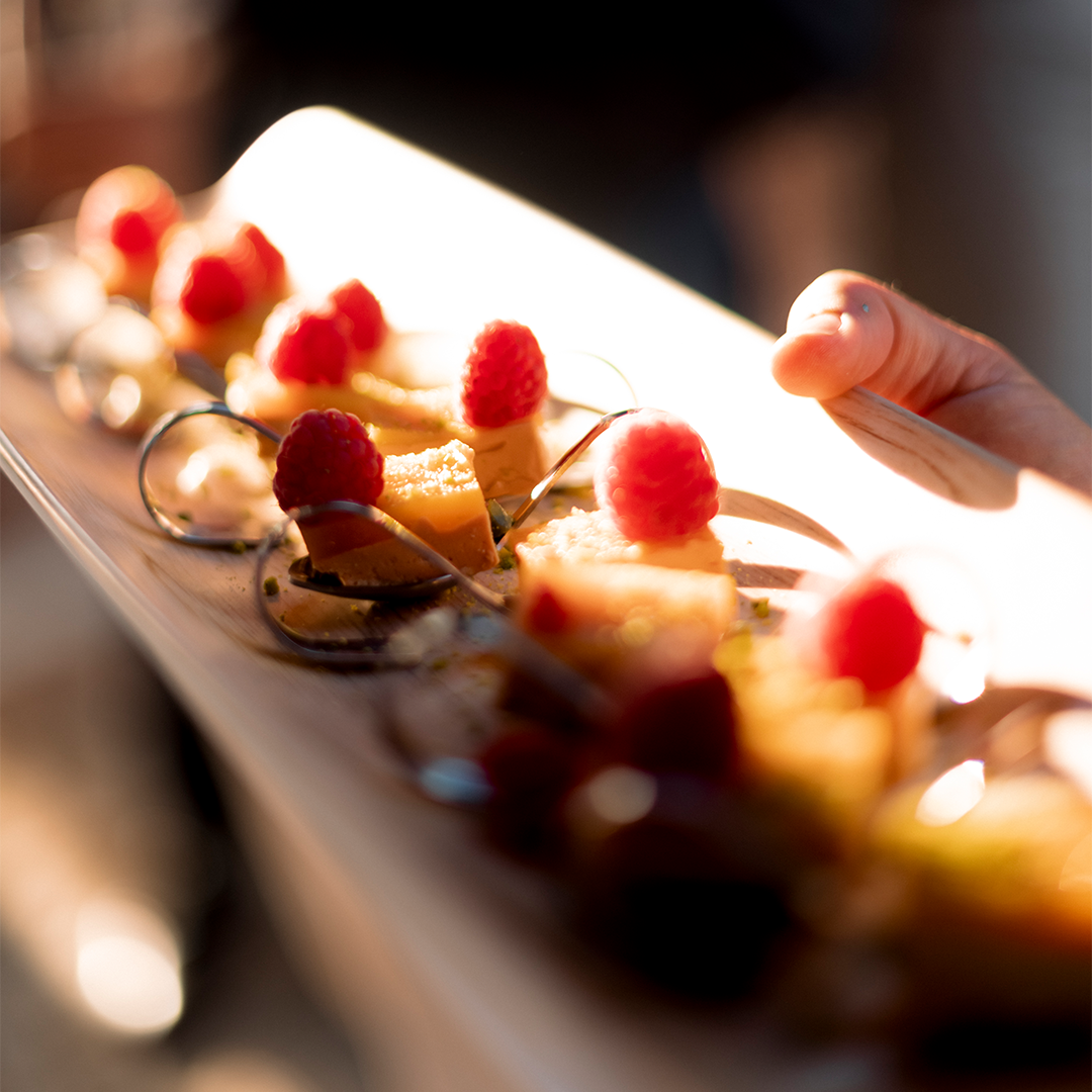 A hand holds a wooden board with neatly arranged appetizers, each topped with a raspberry and garnished with sauce and herbs.