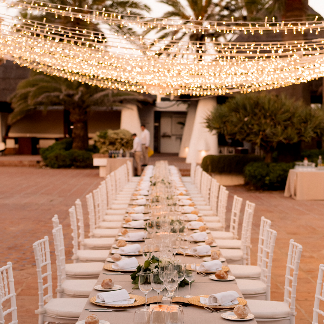 A long outdoor dining table set with white chairs, plates, and glassware, under string lights, with greenery and trees in the background.