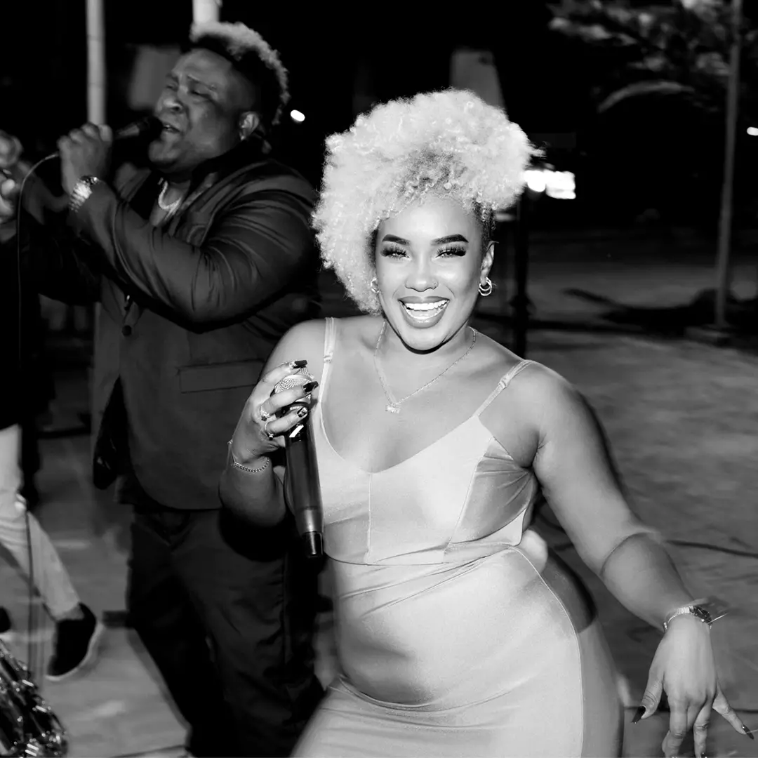 A woman in a sleeveless dress sings and dances with a microphone, while a man in a suit sings behind her on an outdoor stage at night.