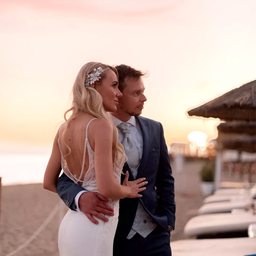 A bride and groom stand close together on a beach at sunset, looking toward the horizon with calm expressions.