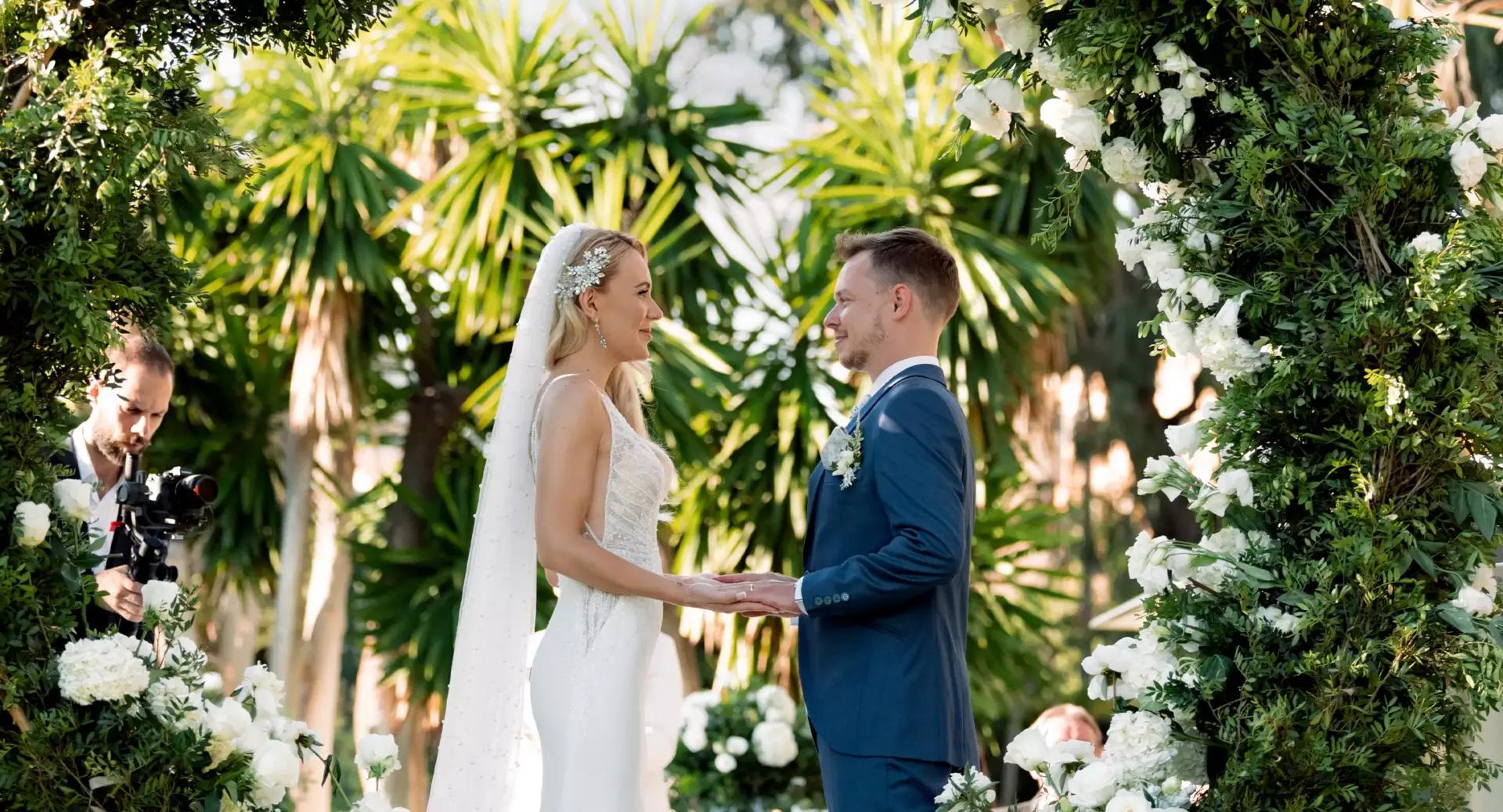 A bride and groom stand facing each other, holding hands during an outdoor wedding ceremony surrounded by greenery and white flowers.