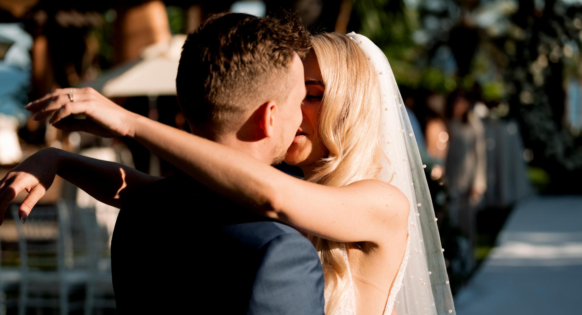 A bride and groom embrace and kiss outdoors on their wedding day, with sunlight highlighting the bride’s veil and hair.