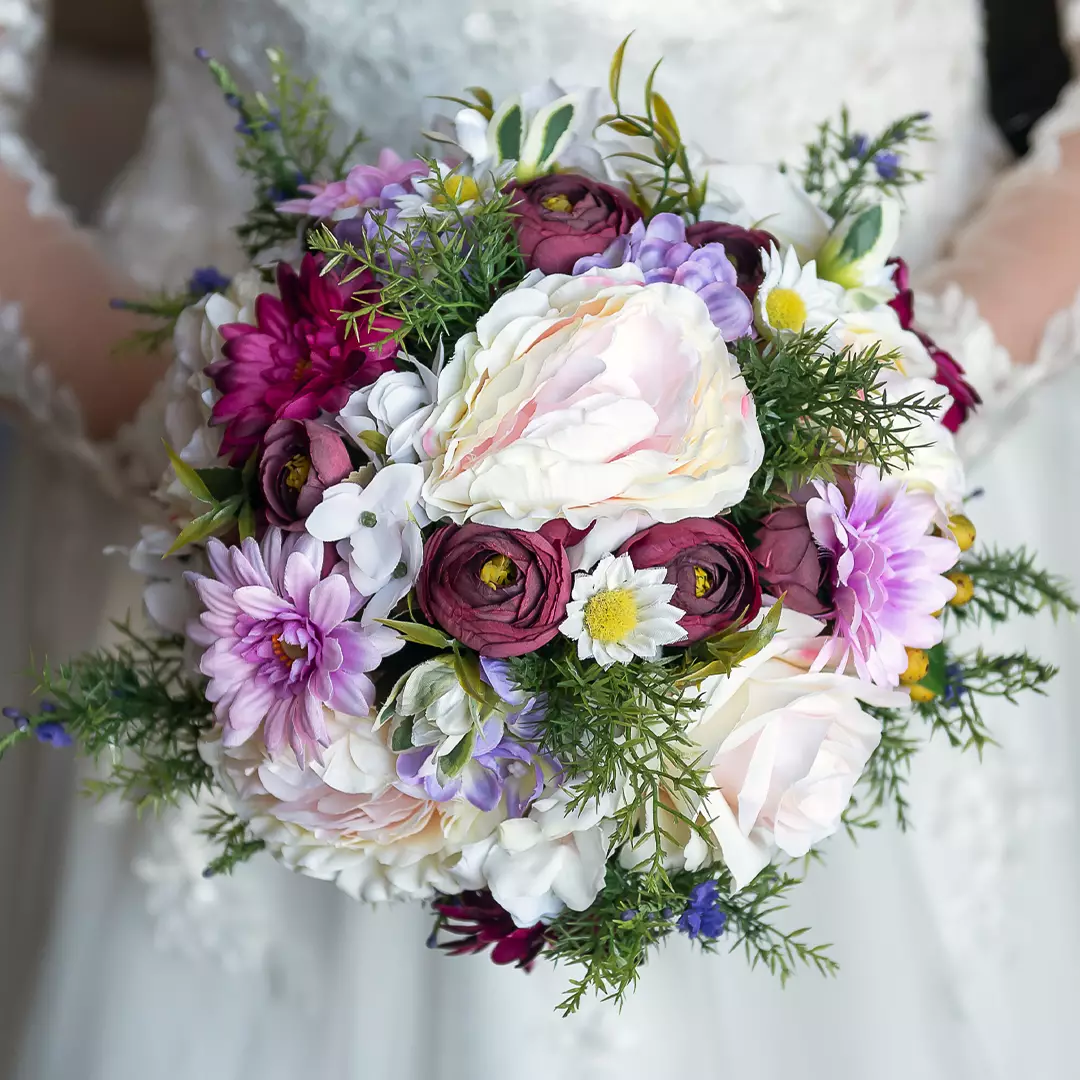 A person in a white lace dress holds a round bouquet of assorted flowers, including white, purple, and burgundy blooms with green foliage.