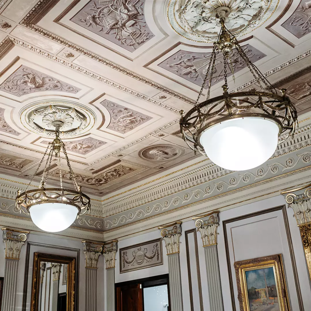Ornate ceiling with decorative molding and two large, round chandeliers in an elegant room with framed artwork and mirrors on the walls.