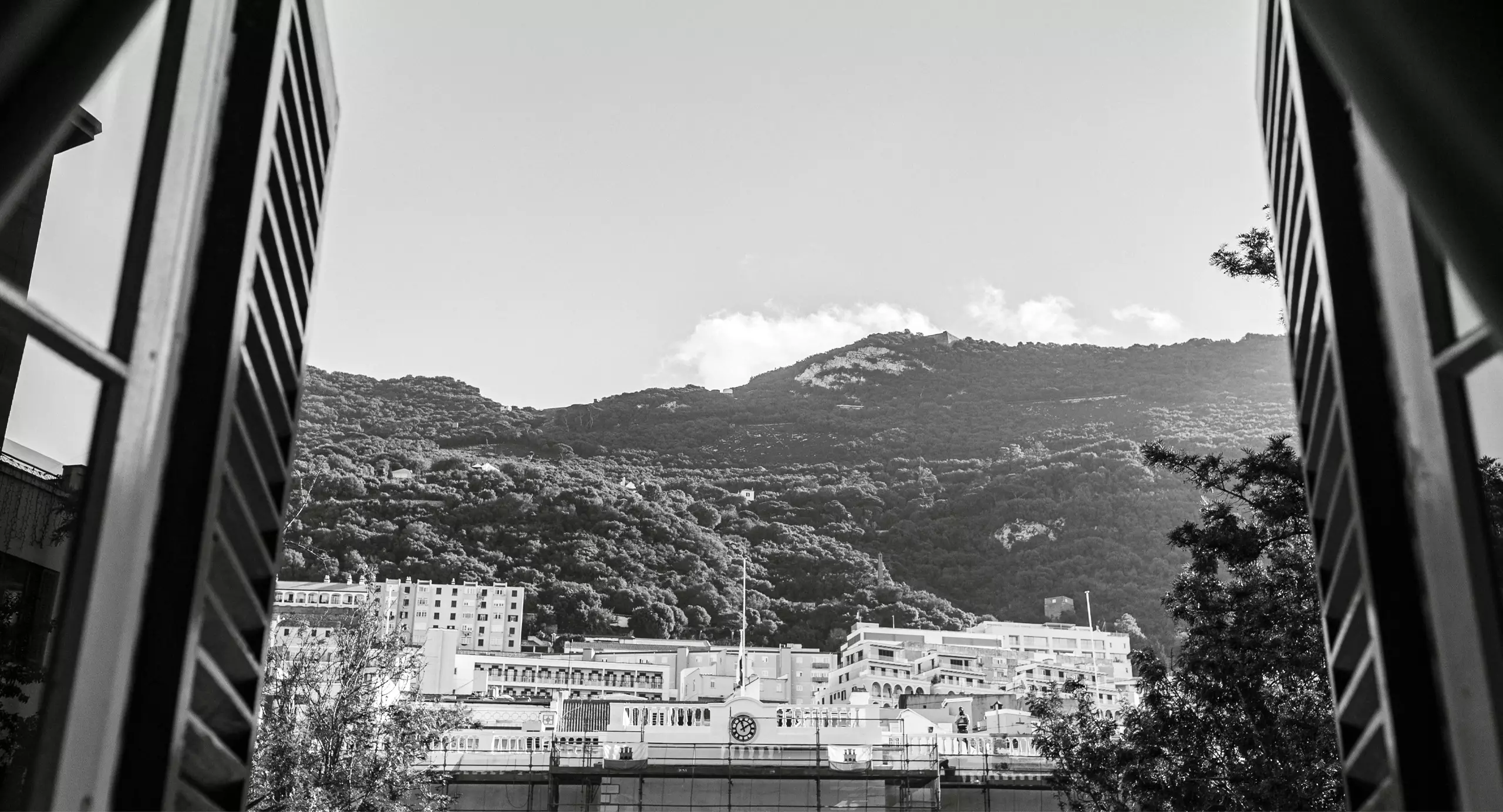 Black-and-white view of buildings and trees at the base of a mountain, seen through an open window with shutters on either side.