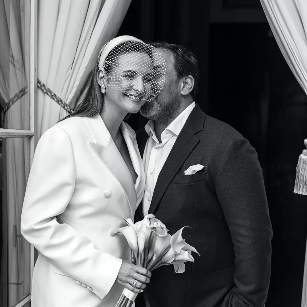 A woman in a white suit and veil holds calla lilies while a man in a dark suit kisses her on the cheek, both standing by a window with curtains.