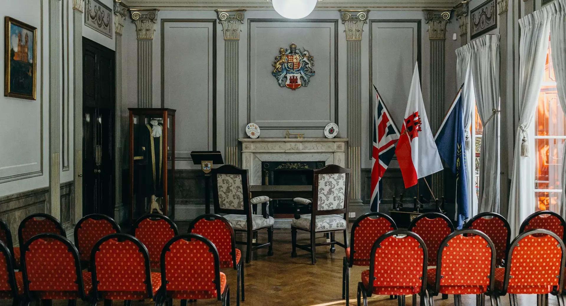 A formal room with red chairs arranged in rows facing a fireplace, British flags, a coat of arms, and two empty chairs at the front.