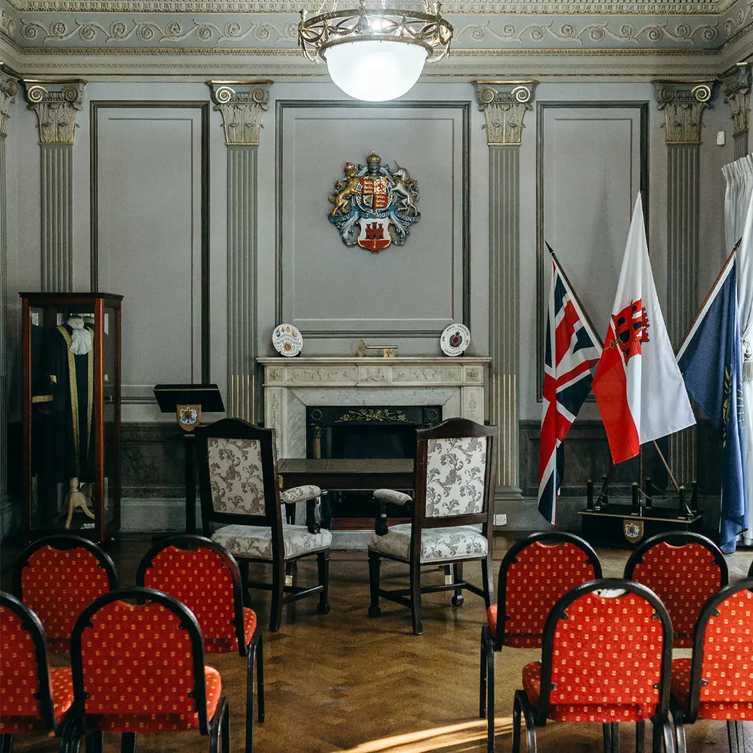 Formal room with red chairs facing two armchairs, a fireplace, display case, flags, and a coat of arms on the wall.