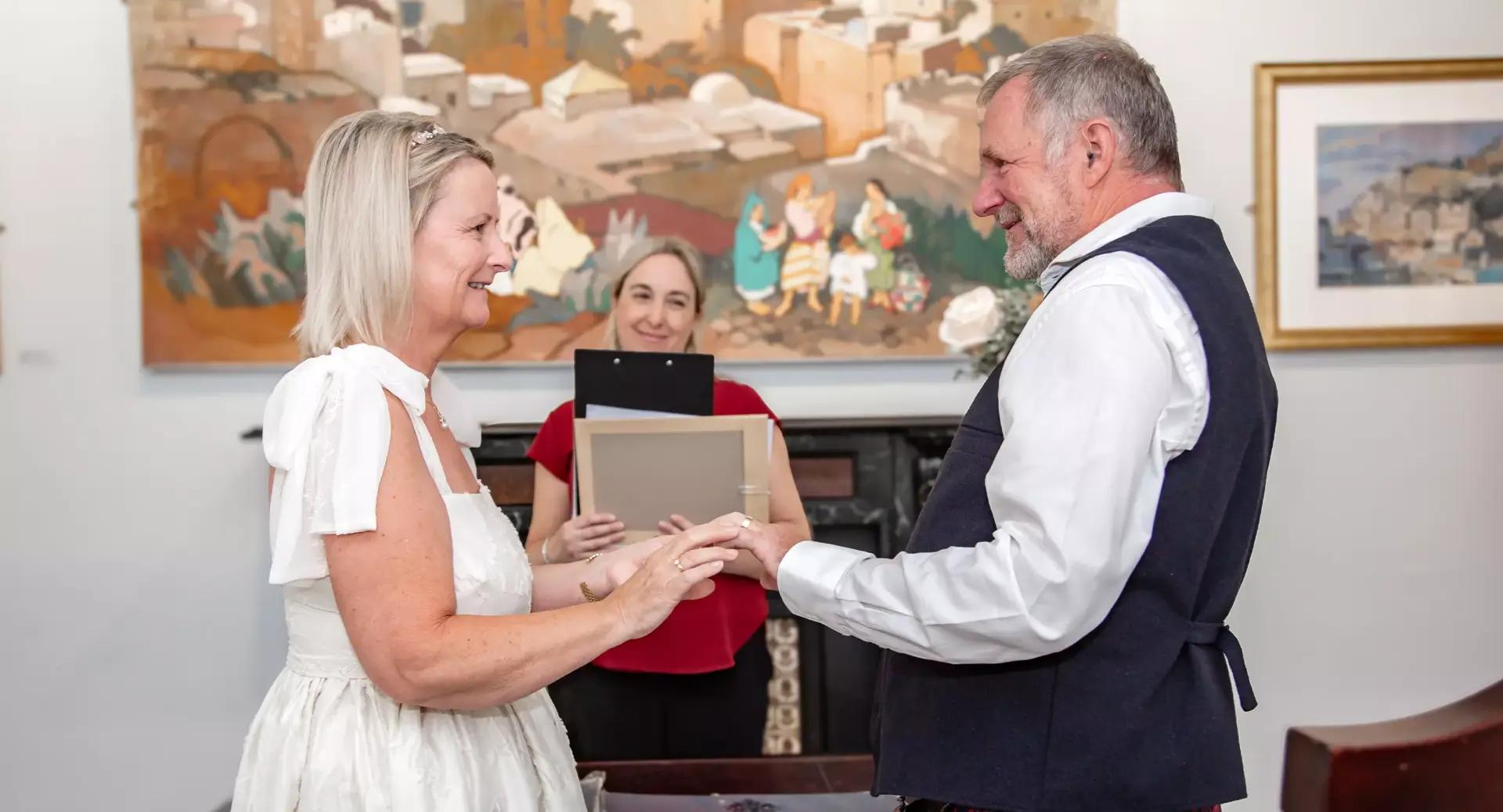 A couple stands facing each other, holding hands and smiling, as a woman officiates their wedding ceremony in an indoor setting.