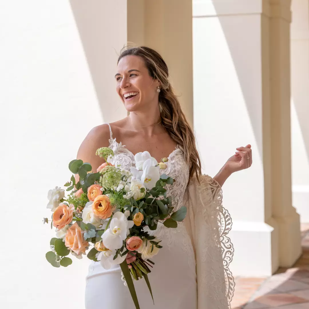 A bride in a white lace wedding dress holds a bouquet of flowers and smiles while standing in a sunlit hallway.