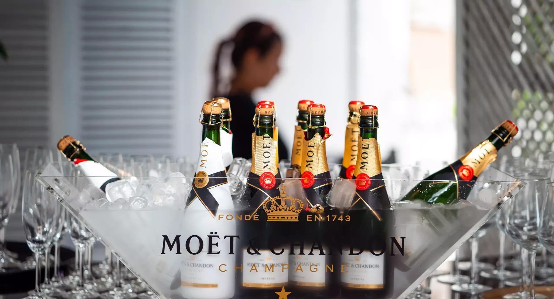 Several bottles of Moët & Chandon champagne are chilled in an ice-filled container, with empty glasses nearby and a blurred person in the background.