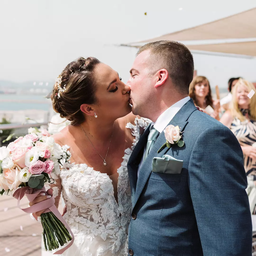 Bride and groom kiss outdoors on their wedding day, with the bride holding a bouquet and guests watching in the background.