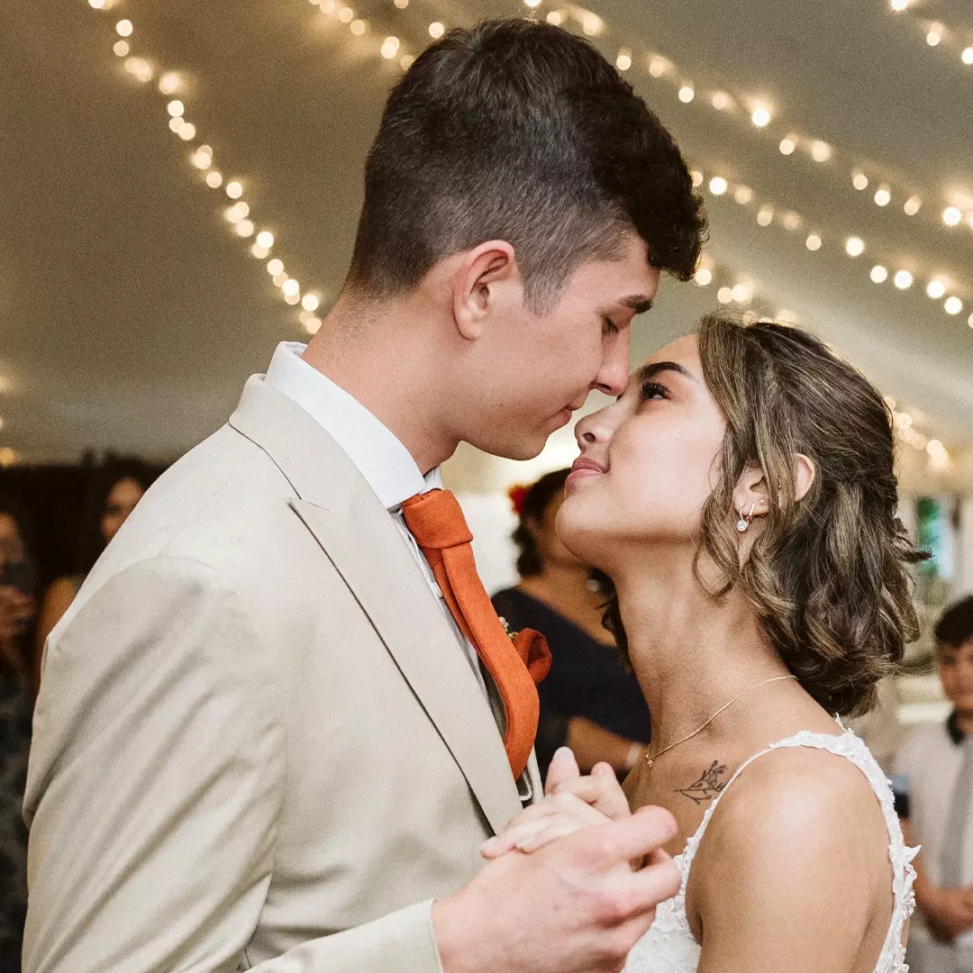 A bride and groom hold hands and gaze at each other while dancing under string lights at a wedding reception.