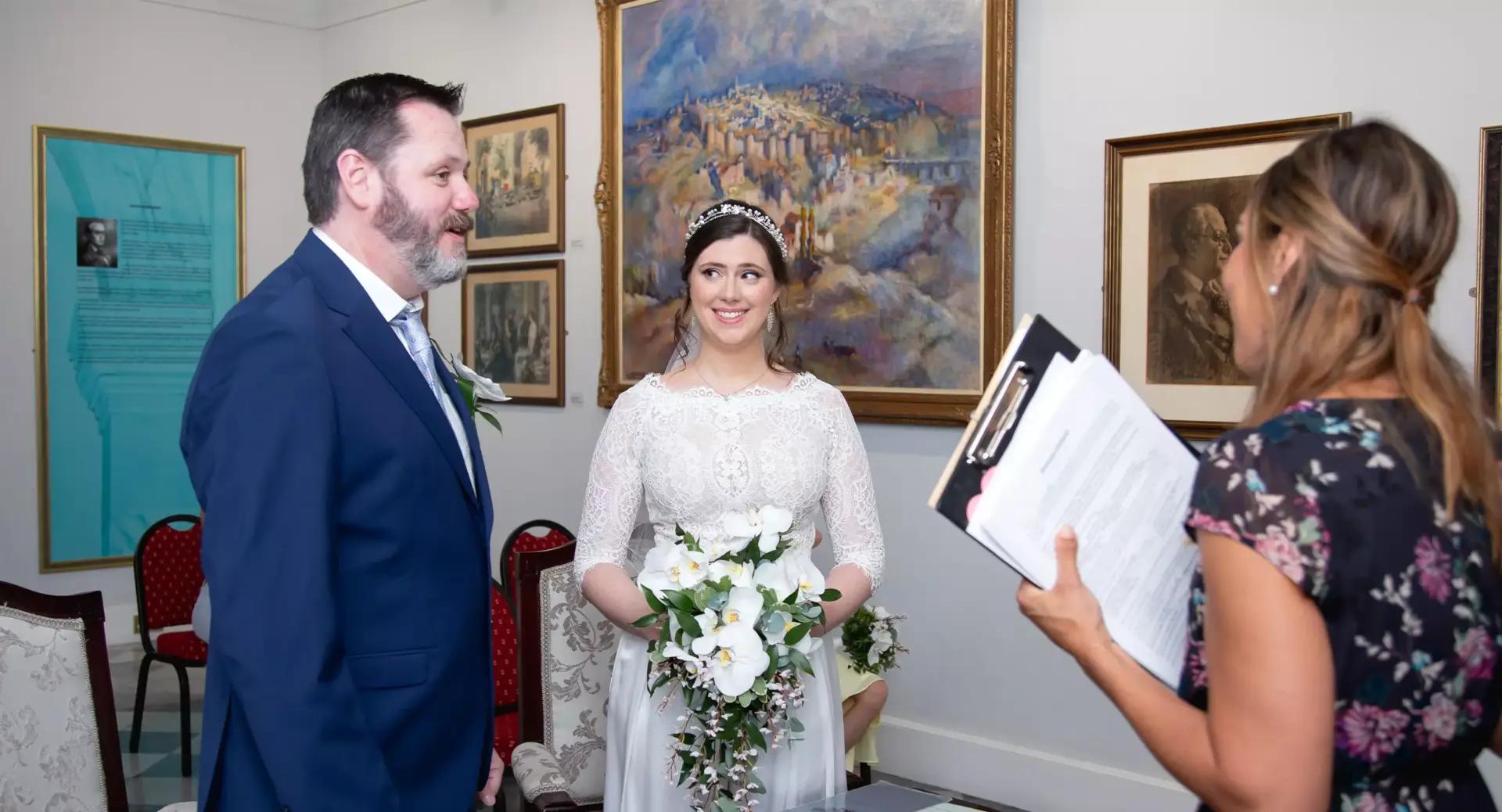 A bride and groom stand together facing a woman holding a clipboard, appearing to officiate their wedding ceremony in an art-filled room.