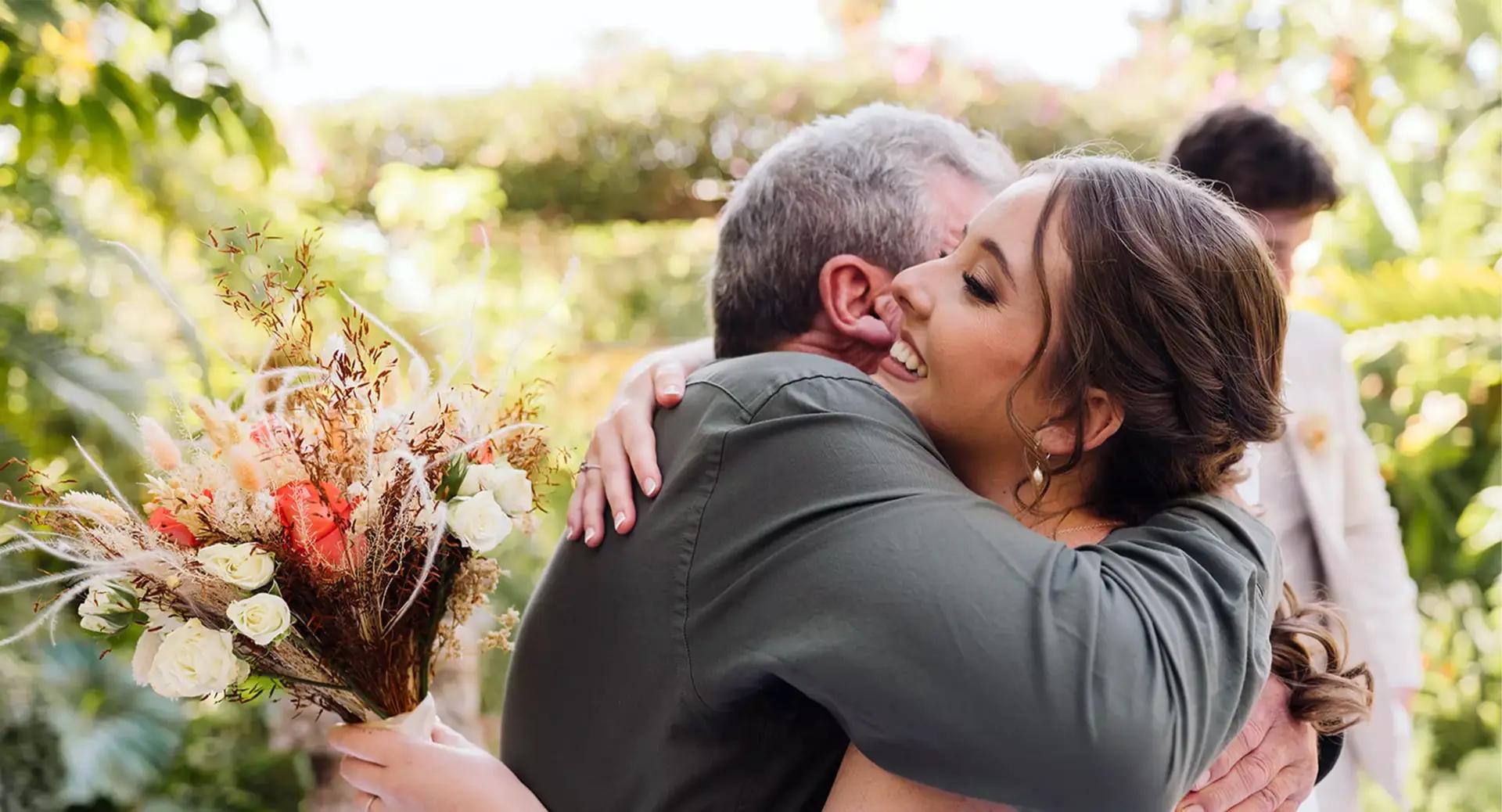 A woman holding a bouquet hugs an older man outdoors, both smiling. Another person stands blurred in the background amid greenery.
