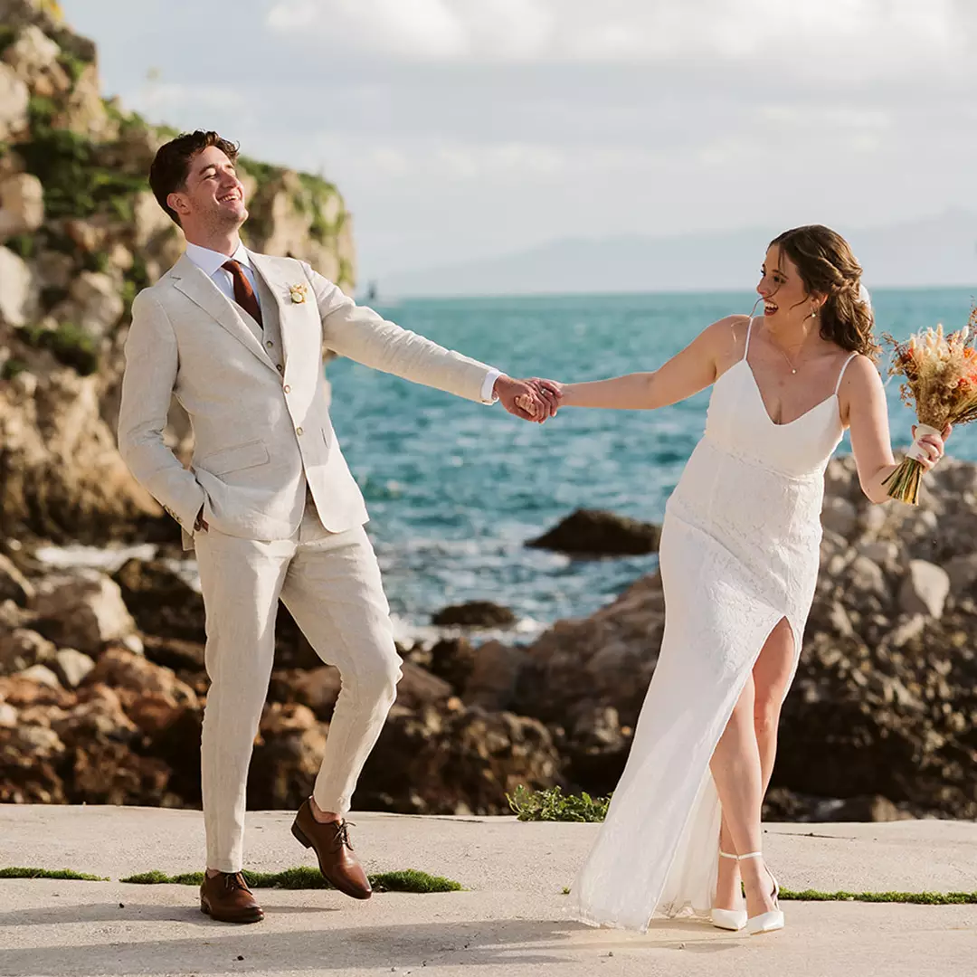 Bride and groom holding hands and smiling by the seaside, with rocky cliffs and blue ocean in the background.