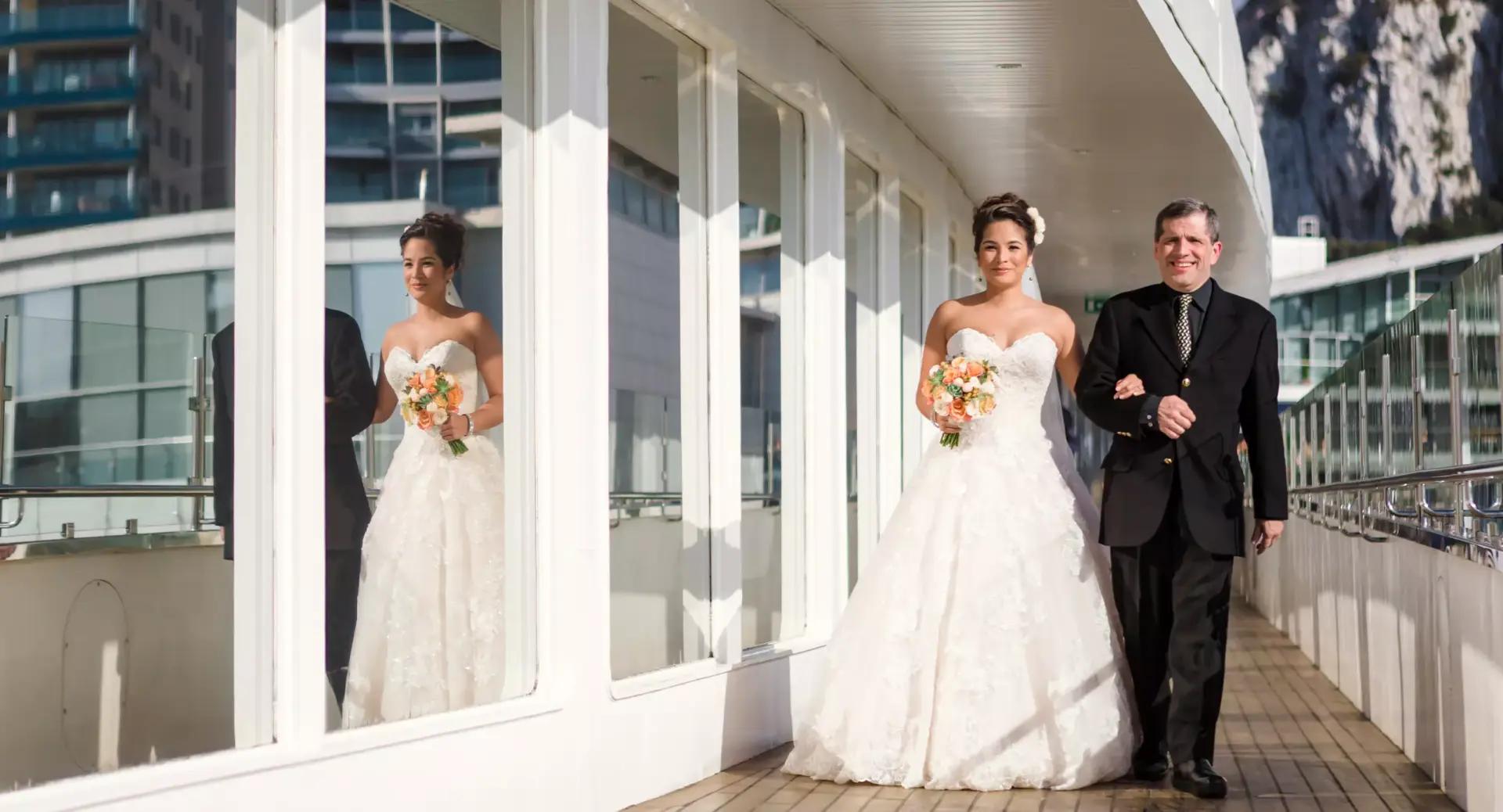 A bride in a white gown holding a bouquet walks arm-in-arm with a man in a suit along a corridor with large windows reflecting their image.