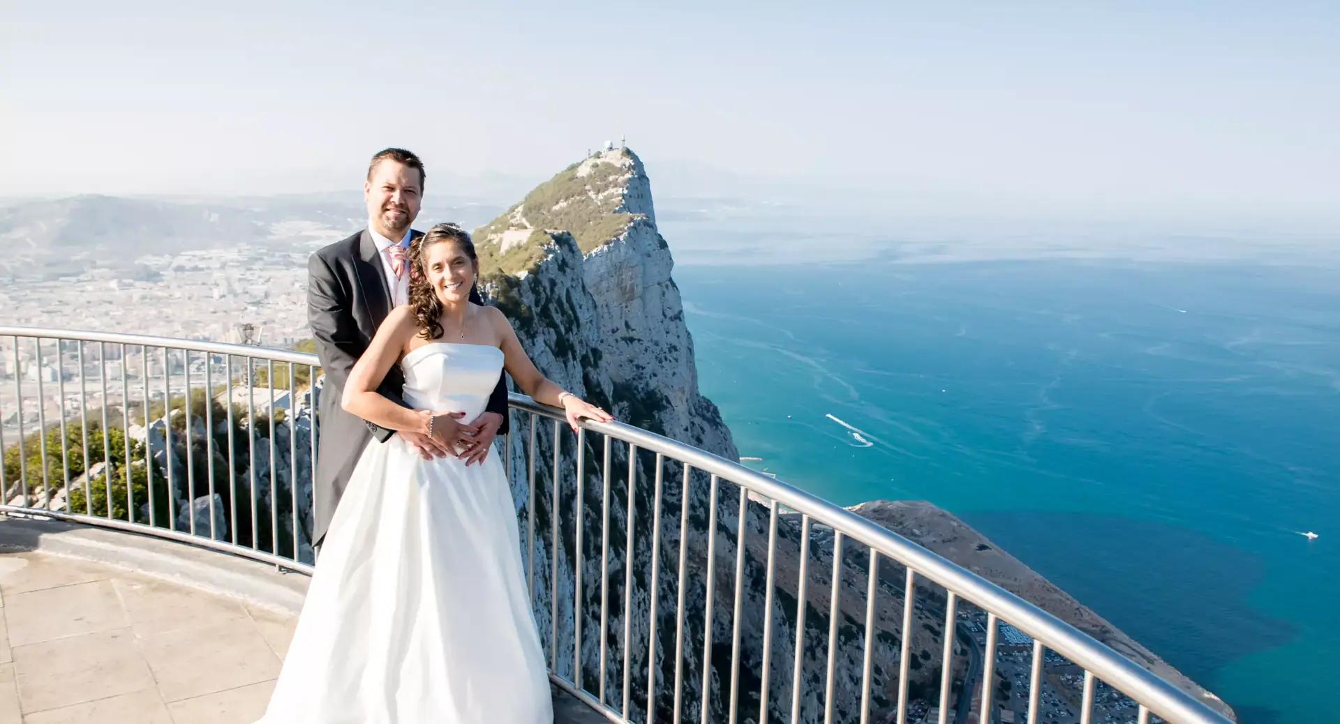 A bride and groom stand together on a balcony overlooking the sea and the Rock of Gibraltar on a clear day.