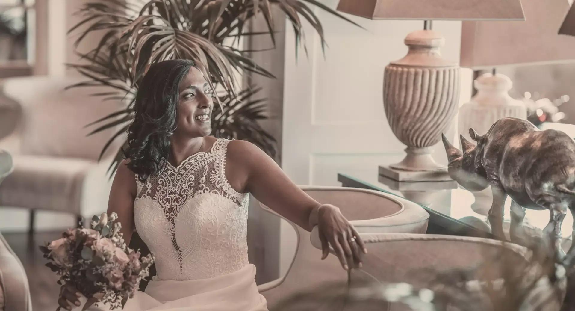 A bride in a lace wedding dress sits indoors holding a bouquet, smiling and reaching toward a rhinoceros sculpture on a table beside her.
