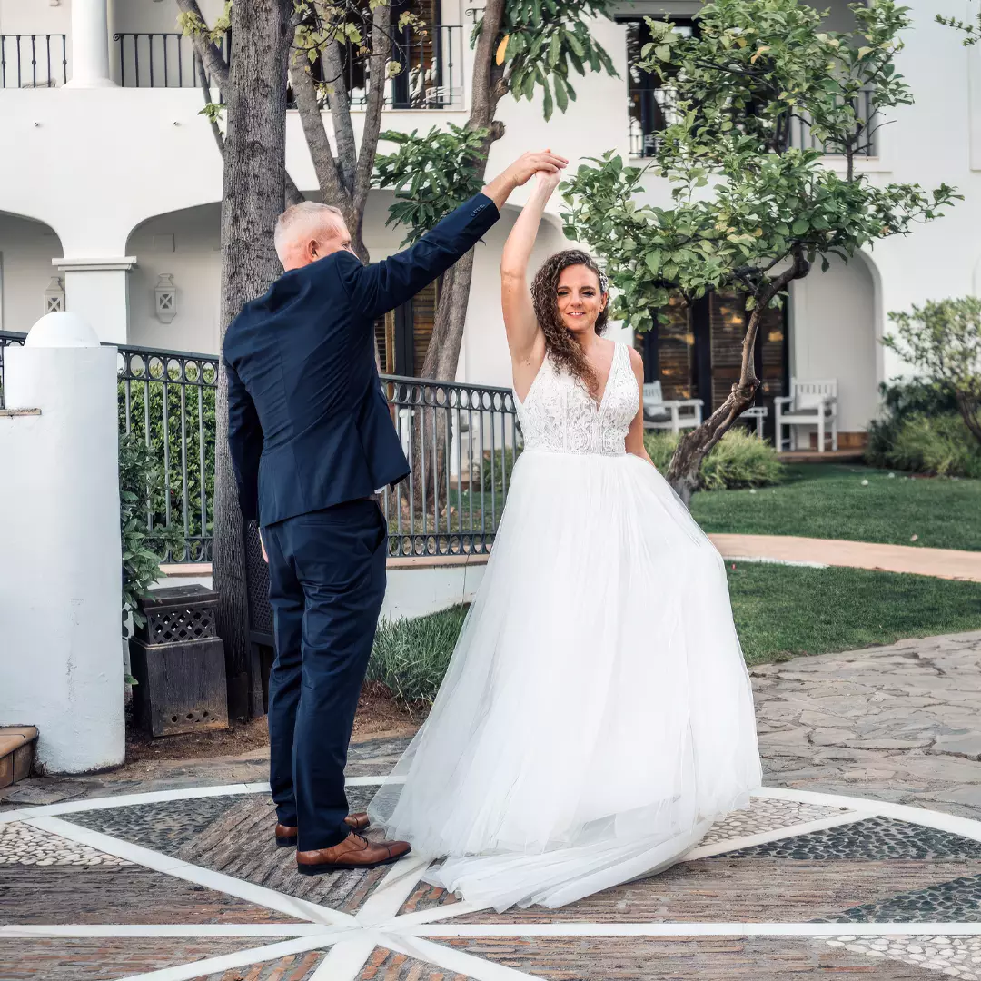 A bride in a white gown twirls with a groom in a dark suit outdoors, standing on a patterned circular stone area near a building with arched windows and greenery.
