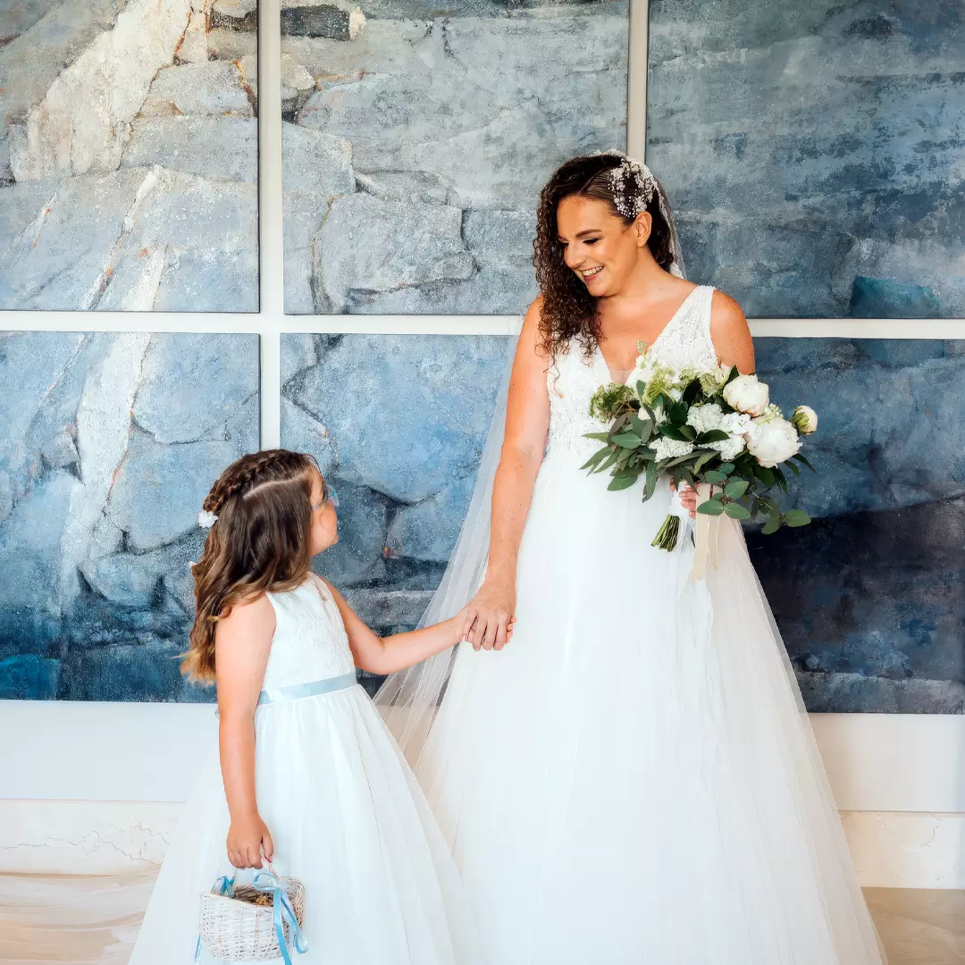A bride in a white dress holds a bouquet and looks at a young girl in a matching dress; they are standing in front of a blue and gray textured wall.