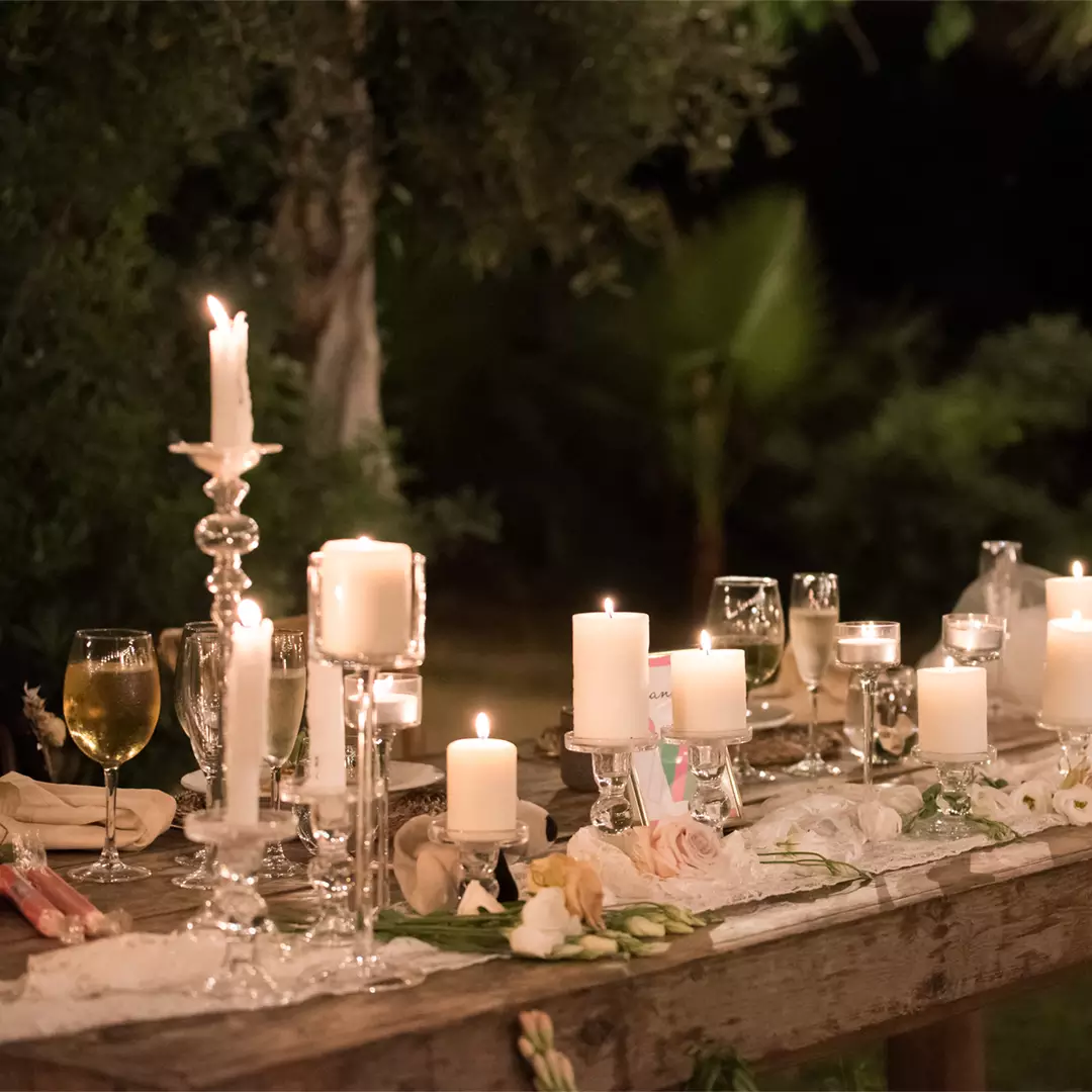 A wooden table set outdoors with lit candles in glass holders, wine glasses, and scattered flowers, creating a warm, elegant ambiance at night.