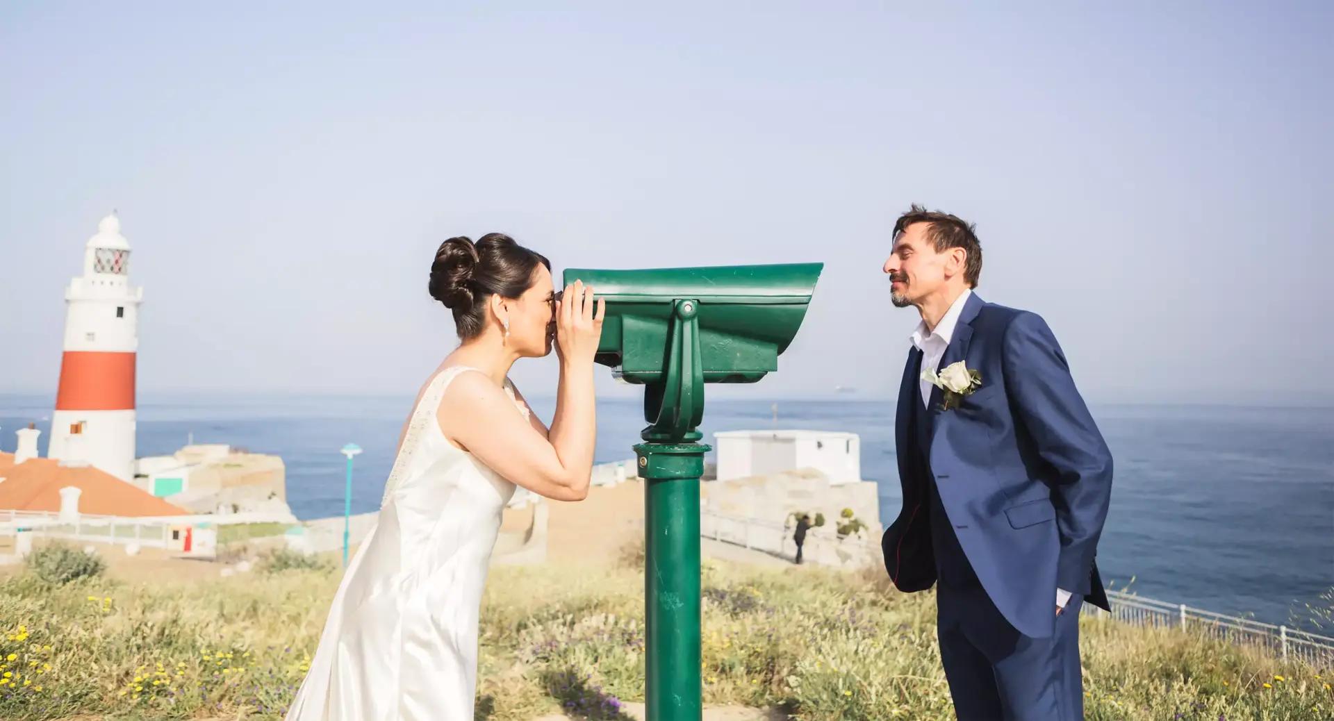 A woman in a white dress looks through a green telescope at a man in a blue suit, with a lighthouse and ocean in the background.