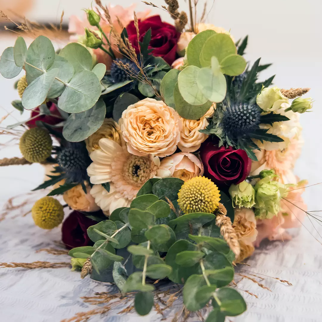 A bouquet of mixed flowers including red roses, pale peach roses, yellow billy balls, white gerbera daisies, eucalyptus leaves, and thistles on a light surface.