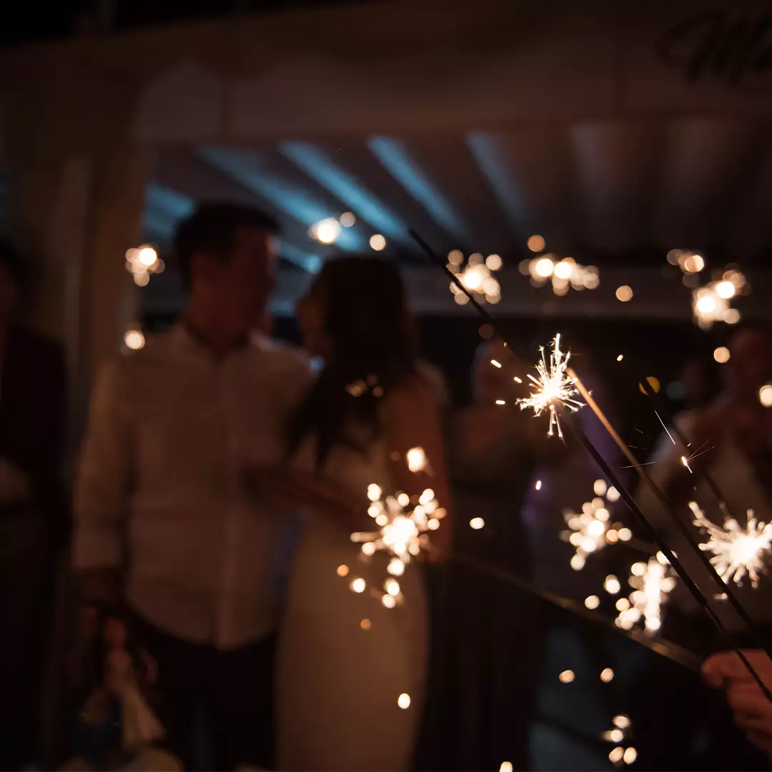 People holding lit sparklers at night, with a couple in the background standing close together and surrounded by others.