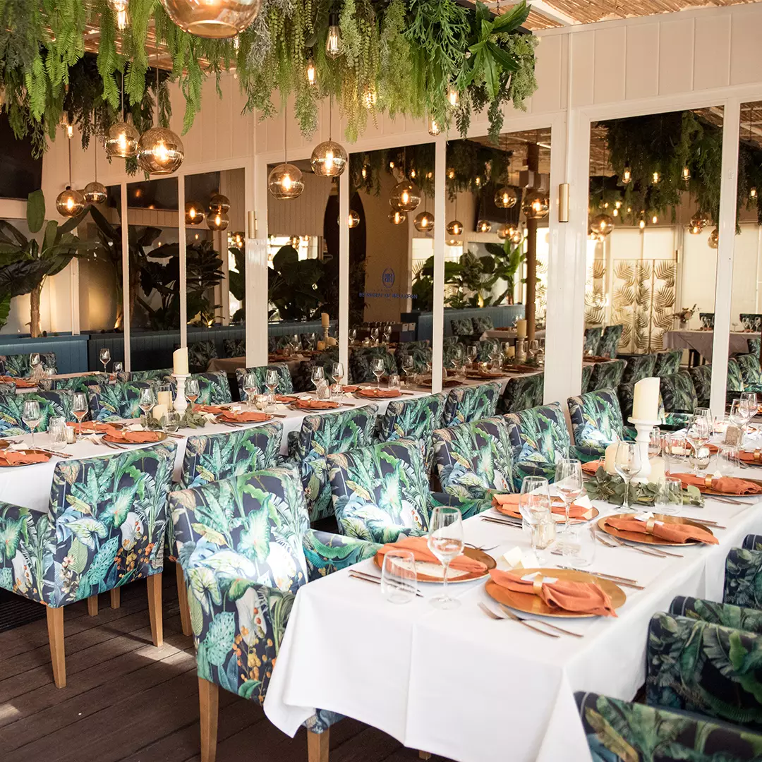 A restaurant dining area with tropical-patterned chairs, white tablecloths, orange napkins, and hanging greenery decorations.