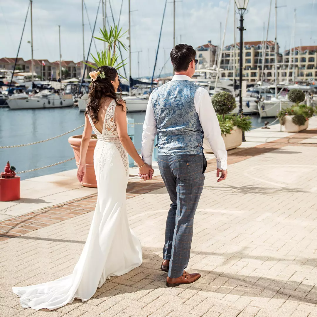 A bride and groom hold hands while walking along a marina with boats docked in the background on a sunny day.